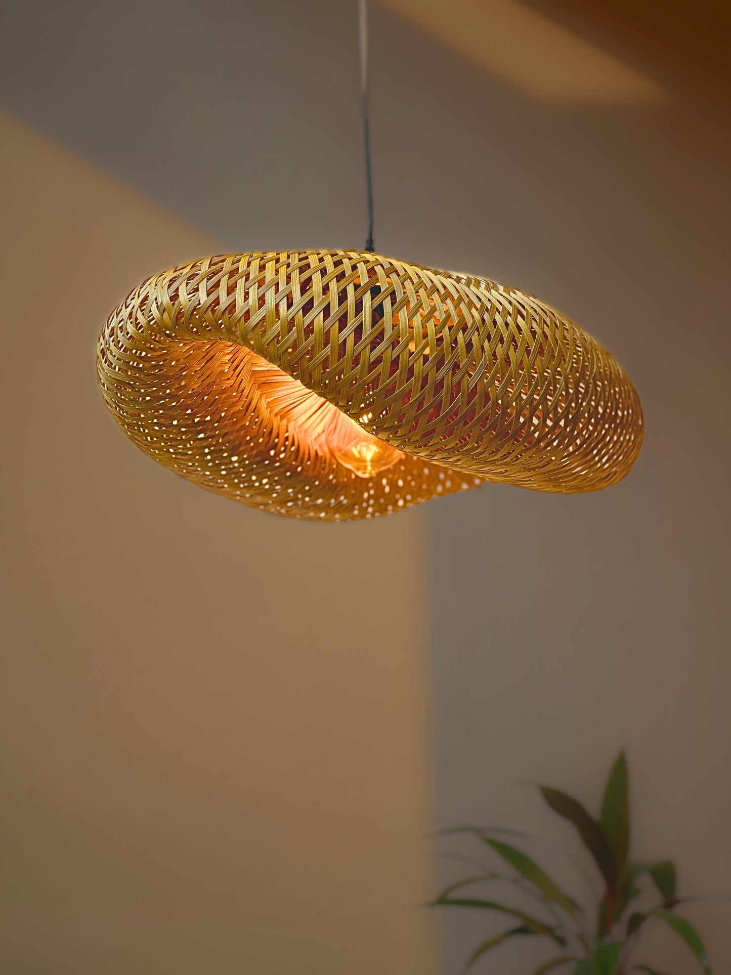 Woven pendant light hanging against a neutral wall with a plant in the corner.